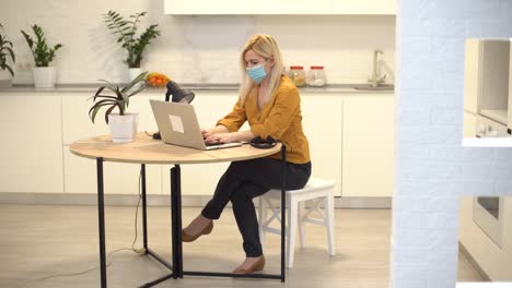 woman-in-protective-mask-works-remotely-on-laptop-at-home-in-kitchen