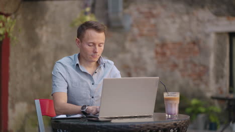 A-young-man-in-a-shirt-is-sitting-at-a-table-with-a-laptop-and-typing-on-the-keyboard.-A-student-can-study-remotely.-A-businessman-conducts-his-business-remotely