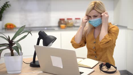 woman-in-protective-mask-works-remotely-on-laptop-at-home-in-the-kitchen,-woman-takes-off-mask