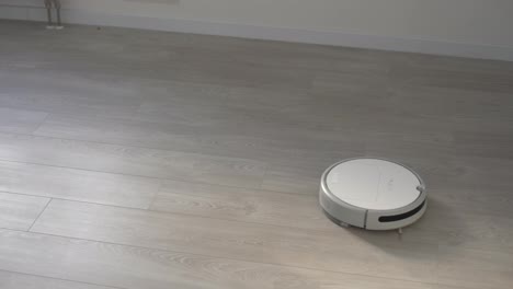 White-robotic-vacuum-cleaner-on-laminate-floor-cleaning-dust-in-living-room-interior.-Smart-housekeeping-technology.