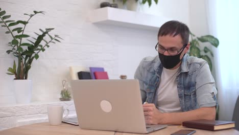 Young-caucasian-man-stay-home-working-from-home-wearing-protective-mask-using-laptop-and-internet.-Cozy-Home-office,-workplace-coronavirus-pandemic,-covid-19-quarantine.-Remote-work,freelancer