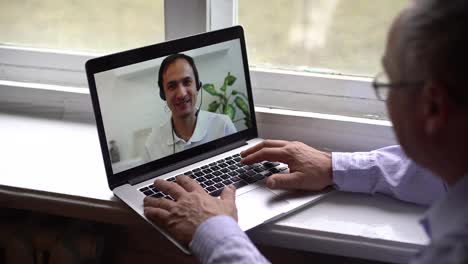 In-Der-Modernen-Medizin-Kommuniziert-Ein-älterer-Mann-Online-Per-Videokommunikation-über-Einen-Laptop-Mit-Dem-Arzt,-Während-Er-Während-Der-Quarantäne-Isolation-Zu-Hause-Am-Tisch-Sitzt
