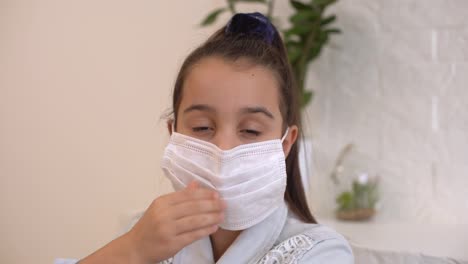 Little-girl-takes-off-her-protective-medical-mask-and-smiles.-Stay-home-mom.-Coronavirus-or-COVID-19.-Cute-little-girl-in-a-protective-mask-at-home-in-a-bright-sunny-room