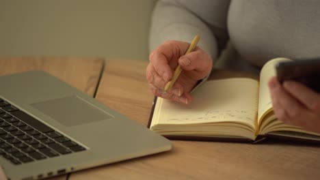 Close-up-wrinkled-male-hands-writing-information.-Old-mature-woman-working-at-office,-using-computer,-handwriting-notes