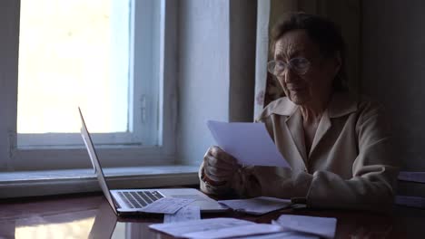 Serious-worried-old-senior-woman-reading-paper-bill-pay-online-at-home,-managing-bank-finances,-calculating-taxes,-fees,-high-rental-rates,-planning-loan-debt-pension-payment-sit-at-kitchen-table.