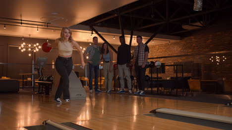 A-young-woman-in-bowling-throws-a-ball-on-the-track-and-knocks-out-a-shot-in-slow-motion-and-jumps-dances-for-joy.-Friends-fans-support-clap.-A-group-of-multi-ethnic-friends-play-bowling-together.