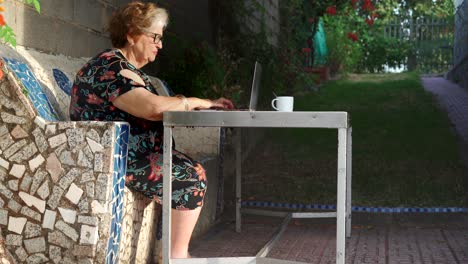 Smiling-aged-female-using-laptop-at-table-in-garden