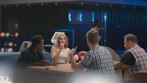 A-multi-ethnic-group-of-friends-in-a-bowling-club-are-chatting-at-the-table-and-smiling-rejoicing-and-high-fiving-a-friend-who-knocked-out-a-strike.-Black-man-and-woman