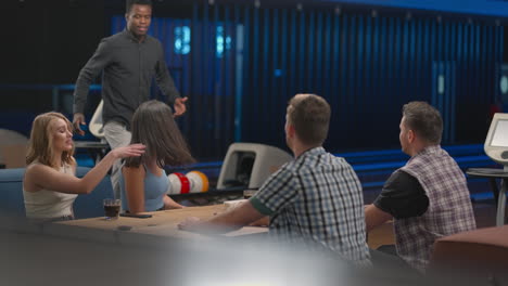A-joyful-black-African-American-man-runs-up-to-friends-giving-five-different-ethnic-groups-of-people-sitting-at-a-table-in-a-bowling-club.-To-celebrate-and-congratulate-on-the-victory-scored-streak.