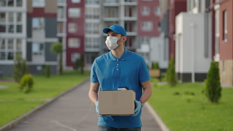 The-male-deliveryman-in-a-cap-and-a-protective-mask-and-gloves-goes-with-a-box-in-his-hands-and-carries-a-parcel-to-the-customer.-Delivering-online-home-orders.