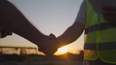 Portrait-of-hands-of-two-builders.-Builder-shaking-hand-the-builder-on-built-house-background.-Close-up-of-a-handshake-of-two-men-in-green-signal-vests-against-the-background-of-the-sun