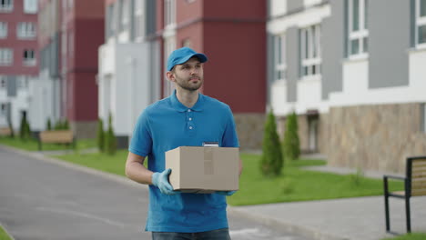 Blue-delivery-man-send-a-package-to-customer-on-before-deliver-cargo.-4k-resolution-and-slow-motion-shot.-Male-postman-with-parcel-in-hands-at-city-street.-Postal-concept.-Delivery-service.