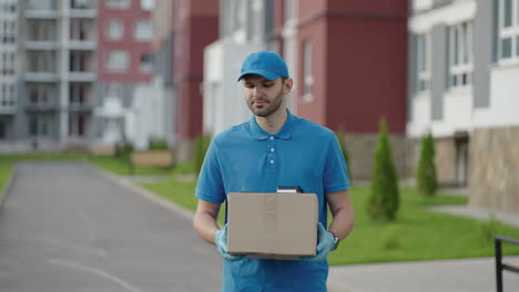 Blue-delivery-man-send-a-package-to-customer-on-before-deliver-cargo.-4k-resolution-and-slow-motion-shot.-Male-postman-with-parcel-in-hands-at-city-street.-Postal-concept.-Delivery-service.