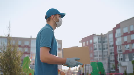 The-male-deliveryman-in-a-cap-and-a-protective-mask-and-gloves-goes-with-a-box-in-his-hands-and-carries-a-parcel-to-the-customer.-Delivering-online-home-orders.