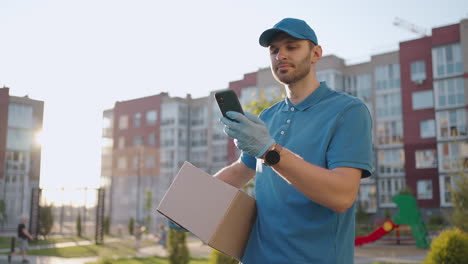 The-postman-with-glasses-carries-the-parcel-and-looks-at-the-delivery-address-via-mobile-phone.-search-for-the-address-of-the-delivery-customer.-Delivery-guy-with-a-cap-and-a-box