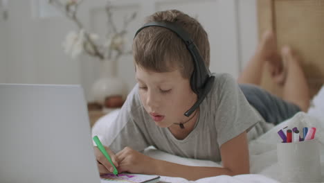 Un-Niño-Pequeño-Está-Sentado-En-El-Sofá-Y-Dibujando-En-Papel-Usando-Una-Computadora-Portátil-Con-Un-Video-Tutorial-Escuchando-Con-Auriculares