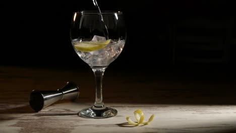 Person-filling-transparent-glass-of-gin-tonic-with-lemon-and-jigger-on-table-in-dark-room