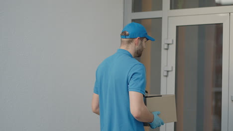 Caucasian-young-pretty-man-delivery-worker-in-blue-cap-walking-the-street-and-carrying-carton-box-while-using-smartphone-looking-for-route.-Male-courier-with-parcel-tapping-and-texting-on-phone.