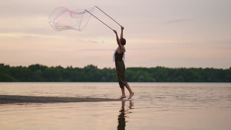 A-young-girl-artist-shows-magic-tricks-using-huge-soap-bubbles.-Create-soap-bubbles-using-sticks-and-rope-at-sunset-to-show-a-theatrical-circus-show