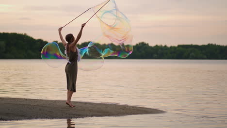 A-young-female-artist-shows-a-soap-bubble-show-blowing-up-huge-soap-bubbles-on-the-shore-of-a-lake-at-sunset.-Show-a-beautiful-show-of-soap-bubbles-in-slow-motion