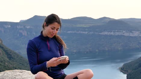 Cheerful-young-ethnic-woman-smiling-and-using-smartphone-on-rocky-cliff