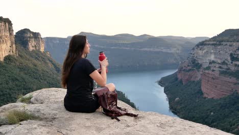 Young-lady-drinking-water-while-recreating-on-rocky-cliff-during-hiking-trip