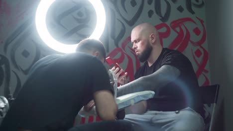 Tattooist-making-tattoo-on-arm-of-client-with-smartphone