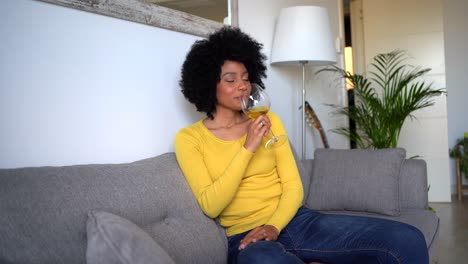 Cheerful-black-woman-sitting-on-sofa-and-tasting-wine-at-home
