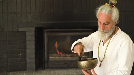 Senior-man-playing-bowl-gong-with-mallet-against-fireplace