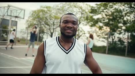 Una-Persona-Negra-Con-Una-Camiseta-Blanca-Posa-Y-Sonríe-En-La-Cancha-De-Baloncesto-Frente-A-Sus-Amigos-Que-Juegan-Baloncesto