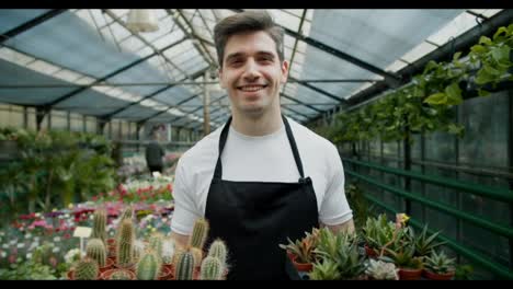 A-young-employee-of-a-specialized-store-selling-flowers-holds-flowers-and-cacti-in-their-hands-in-pots-and-looks-at-the-camera