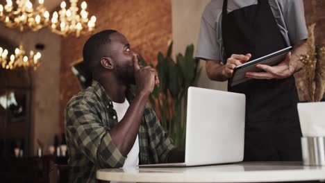 A-black-person-with-a-beard-works-on-a-laptop-in-a-cafe,-a-waiter-approaches-them-and-takes-an-order