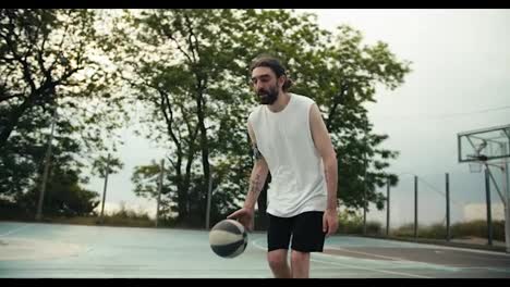A-man-in-a-white-t-shirt-and-black-shorts-skillfully-dribbles-a-basketball-on-a-basketball-court-outside-in-summer.-Active-leisure-sports-outdoor