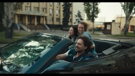 A-happy-trio:-a-guy-and-two-girls-are-riding-in-a-gray-convertible.-A-blonde-girl-and-a-brunette-girl-have-fun-and-rejoice.-Happy-cabriolet-ride