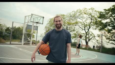 Nahaufnahme-Eines-Rothaarigen-Mannes-In-Einem-Grauen-T-Shirt-Mit-Einem-Basketball-In-Der-Hand,-Der-Vor-Dem-Hintergrund-Einer-Mannschaft-Posiert,-Die-Draußen-Auf-Dem-Basketballplatz-Basketball-Spielt