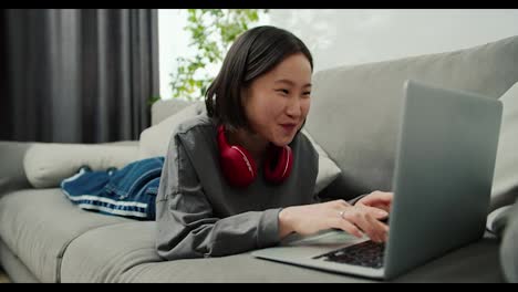 Excited-asian-woman-typing-on-laptop-while-laying-on-sofa,-communicating-with-friends