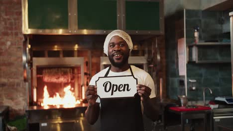 A-Black-person-in-a-special-headdress-poses-with-an-Open-sign-against-the-background-of-a-barbecue-in-a-doner-market