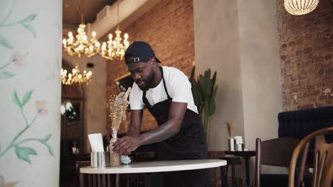 A-Black-person-in-a-black-apron-and-white-T-shirt-wipes-the-table-with-a-special-towel-in-a-cafe.-High-quality-video
