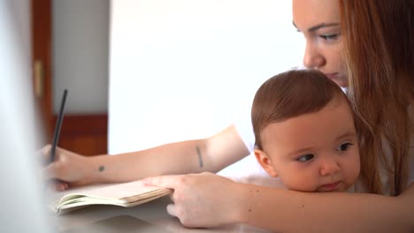 Young-mother-hugging-baby-on-laps-and-writing-in-copybook