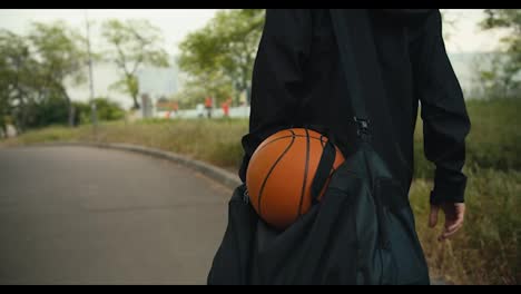 Close-up-shot-of-a-man-in-a-black-jacket-and-with-a-black-backpack-goes-to-a-basketball-game-with-a-basketball-down-the-street