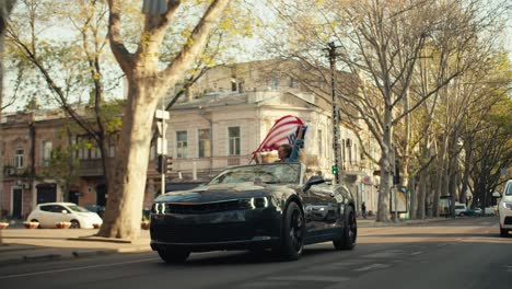 Dark-gray-glossy-convertible-rides-along-a-sunny-street-a-girl-who-sits-in-a-convertible-waving-the-flag-of-the-United-States-of-America