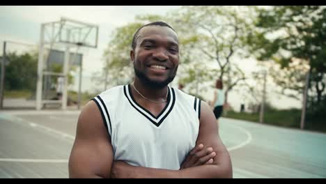 A-Black-man-in-a-white-t-shirt-poses-in-front-of-his-friends-who-play-basketball-outdoor-on-the-court-in-summer