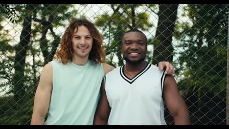 Portrait-of-two-friends-red-haired-curly-haired-man-and-a-Black-man-in-a-white-t-shirt-posing-and-looking-at-the-camera-and-smiling-against-the-background-of-a-mesh-fence-on-a-bucksball-court