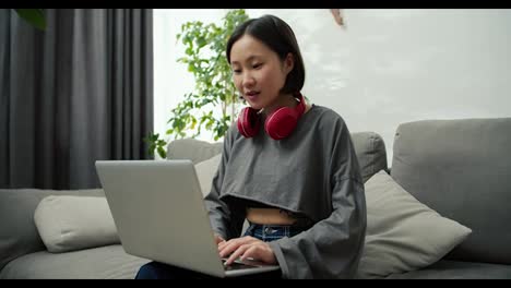 Junge-Asiatische-Frau-Tippt-In-Lässiger-Kleidung-Auf-Dem-Laptop-Und-Arbeitet-Zu-Hause-Auf-Dem-Bequemen-Sofa-Im-Wohnzimmer