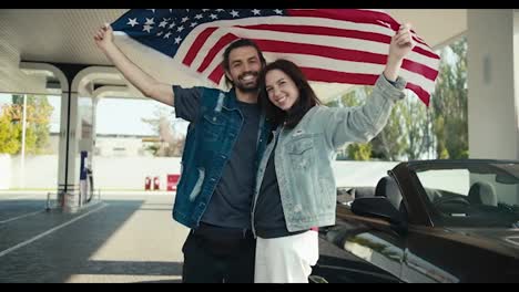 A-happy-couple-of-a-guy-and-a-brunette-girl-in-leather-jackets-raise-the-US-flag-above-their-heads,-they-stand-near-a-gray