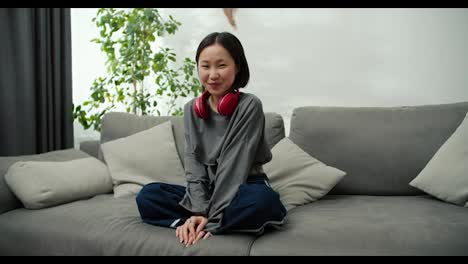 Portrait-of-gentle-asian-girl-wearing-wireless-headphones,-sitting-on-couch-and-smiling-to-the-camera