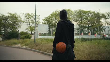 Rear-view-of-a-brunette-man-in-a-black-jacket-with-a-black-backpack-and-a-basketball-goes-to-a-basketball-game-on-a-street-with-a-lot-of-trees