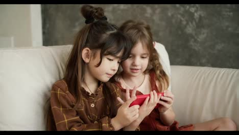 Two-smiling-children-sit-on-couch-look-at-cellphone-screen,-have-fun-together