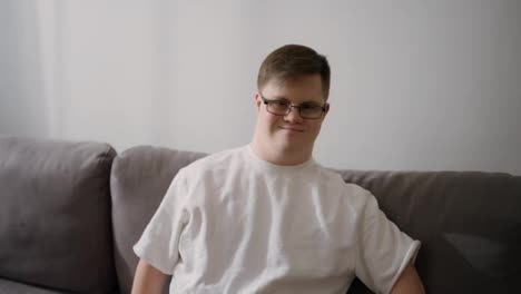 Portrait-of-happy-caucasian-man-in-white-T-shirt-with-Down-Syndrome-sitting-on-couch-at-home