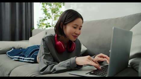 Smiling-asian-woman-typing-on-laptop-while-laying-on-sofa,-working-in-living-room-at-home